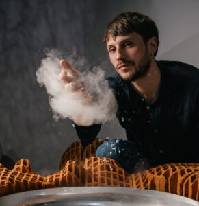 Portrait de l'artiste Arthur Hoffner devant son oeuvre dans l’exposition CHAOSMOS au MAIF Social Club, invitation faite à 12 artistes de nous raconter le cosmos et d’inventer de nouvelles manières d’habiter la Terre, par la commissaire Jos Auzende et scénographiée par Lucie Gautrain, à Paris.